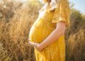 schwangere Frau, die gelbes Blumenkleid trägt, das steht, während sie ihren Bauch berührt und ihrer rechten Seite nahe dem braunen Feld während des Tages gegenübersteht