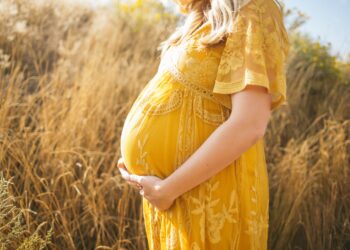 妊娠中の女性は彼女のおなかに触れながら昼間茶色のフィールドの近くの彼女の右側に直面しながら立っている黄色の花柄のドレスを着ています。