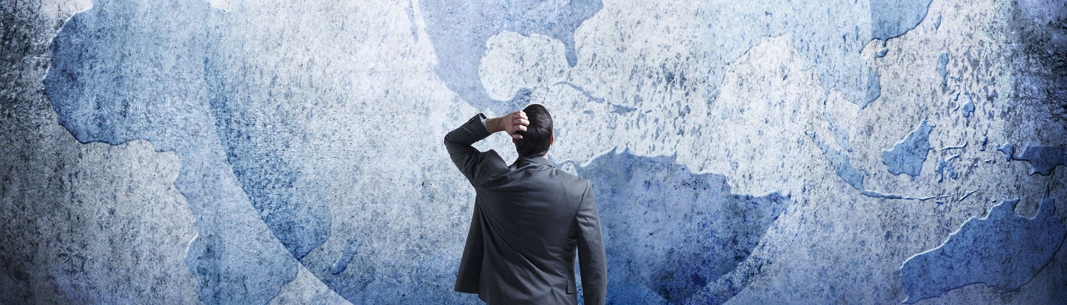 A rear view of a businessman as he stands and looks up at three interlocking globes on the wall in front of him as he tries to sort out the mess of geopolitical events. Map source material courtesy of https://images.nasa.gov/