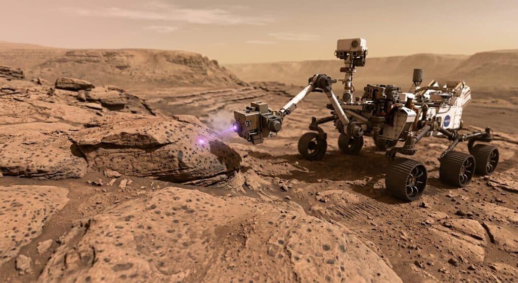A photorealistic, cinematic close-up of the Martian surface inside Jezero Crater, showing ancient reddish-brown mudstone rocks with subtle dark leopard-like spots, illuminated by the pale pinkish Martian sunlight. The NASA Perseverance rover is visible in the mid-ground, its SHERLOC instrument arm extended toward a natural rock surface, emitting a faint ultraviolet laser beam. The terrain features layered sedimentary formations along an ancient dried riverbed. The atmosphere is dusty and amber-tinted, with a hazy horizon. The mood is one of quiet scientific discovery — vast, mysterious, and awe-inspiring. Editorial photography style, shallow depth of field, dramatic directional lighting.