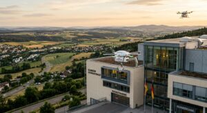 A small white medical delivery drone hovers precisely over a modern hospital building in Germany, gently docking onto a metallic hook mounted above a large window. The scene is set at golden hour, with soft northern European light casting warm tones over the clinical architecture. Below, a sprawling rural landscape stretches into the distance, dotted with fields and small towns connected by winding roads. In the background sky, another drone is visible in flight, carrying a small payload container. The mood is quietly futuristic yet grounded — technological innovation integrated seamlessly into everyday healthcare infrastructure. The composition emphasizes the contrast between the high-tech drone and the traditional hospital building, conveying urgency, precision, and the promise of faster emergency response. Photorealistic editorial style, wide-angle perspective, dramatic depth of field.