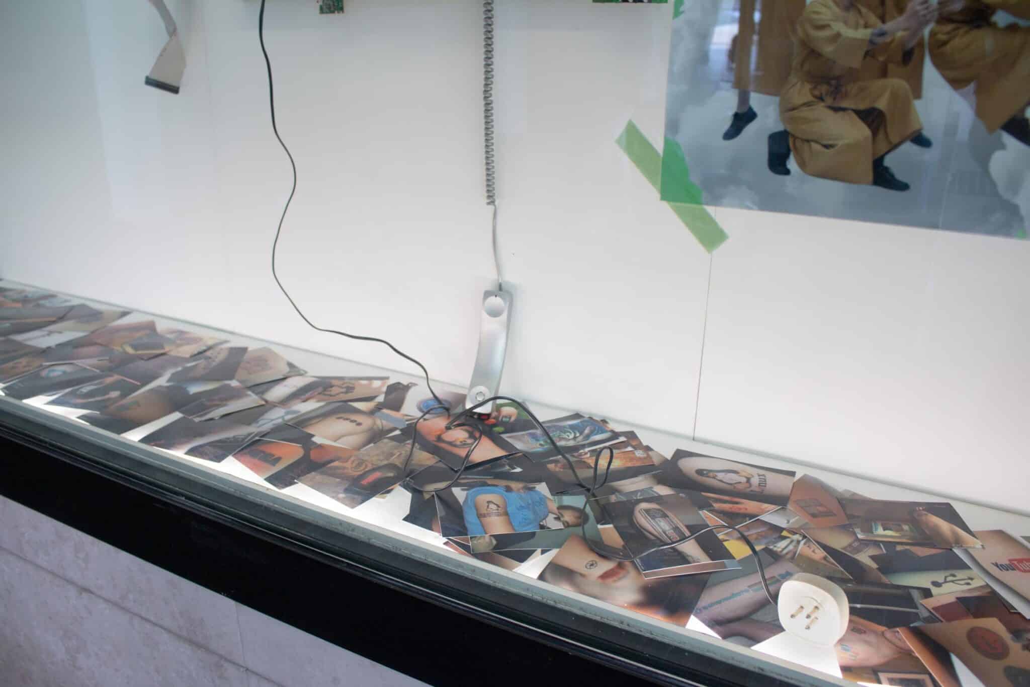 Decine di fotografie di tatuaggi di loghi tecnologici sul piano di una vetrina dell'installazione Atrophia, con una cornetta telefonica dal filo a spirale e una spina bianca
