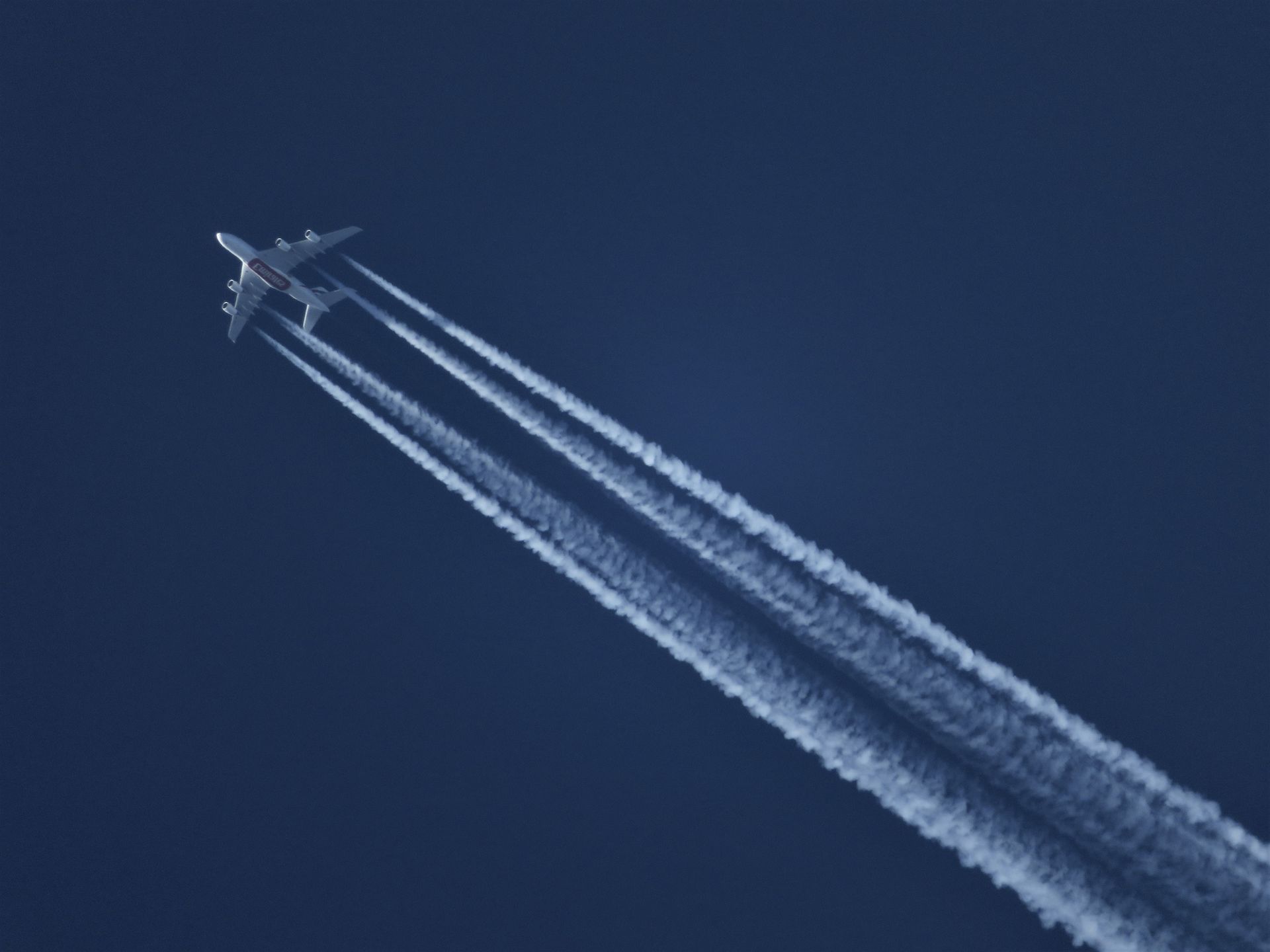 Un grande aereo a quattro motori in quota lascia quattro lunghe scie bianche in un cielo blu scuro. Contrails