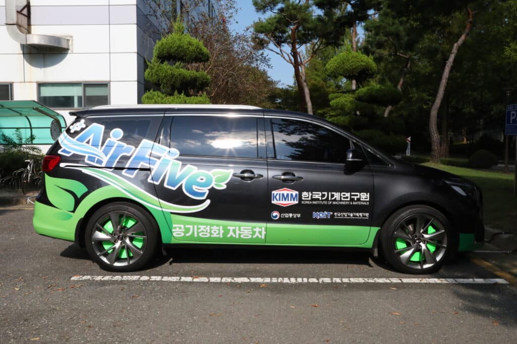 Una Kia Carnival nera e verde con la scritta AirFive e il logo KIMM, parcheggiata accanto a un edificio. Polveri sottili