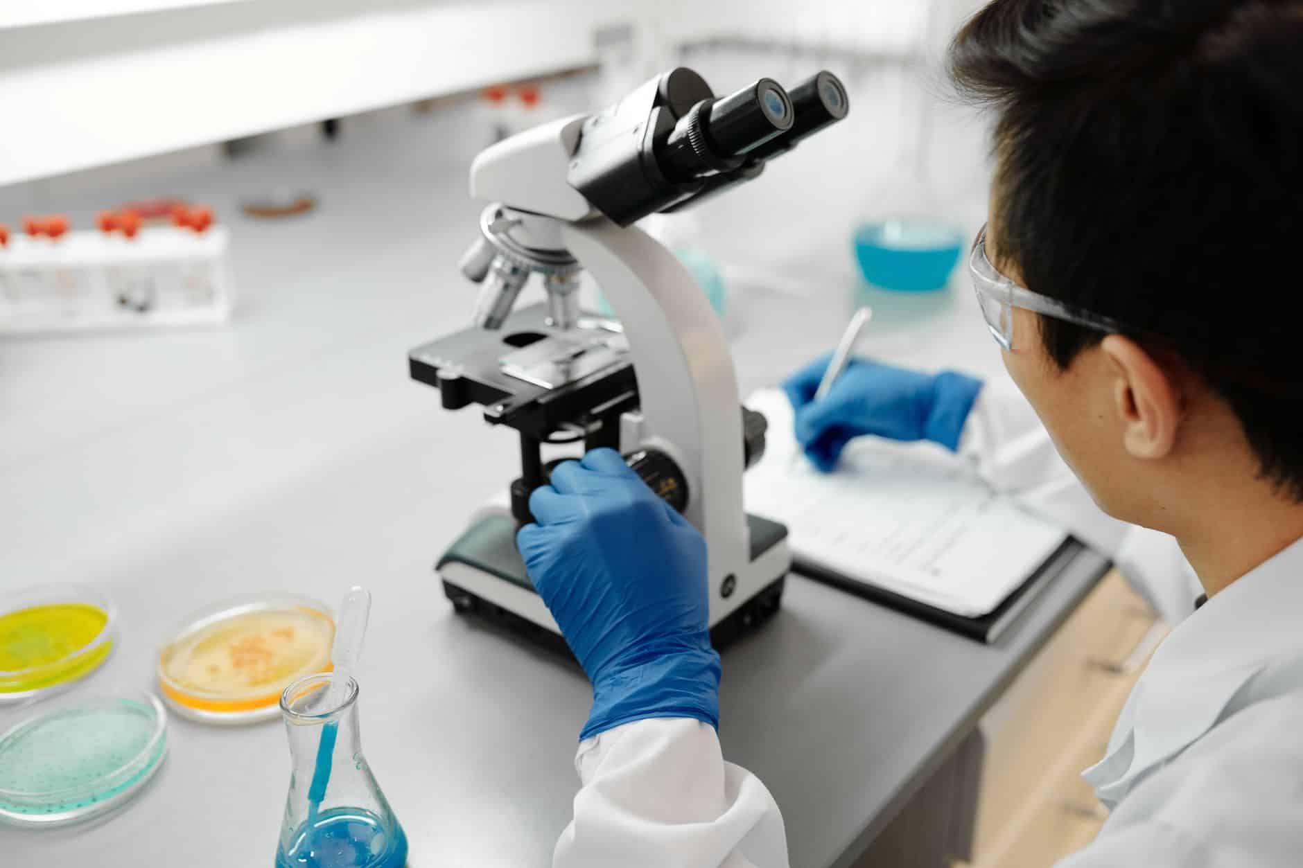 Un ricercatore con occhiali protettivi e guanti blu regola un microscopio, con capsule di Petri sul banco. Cancro al colon