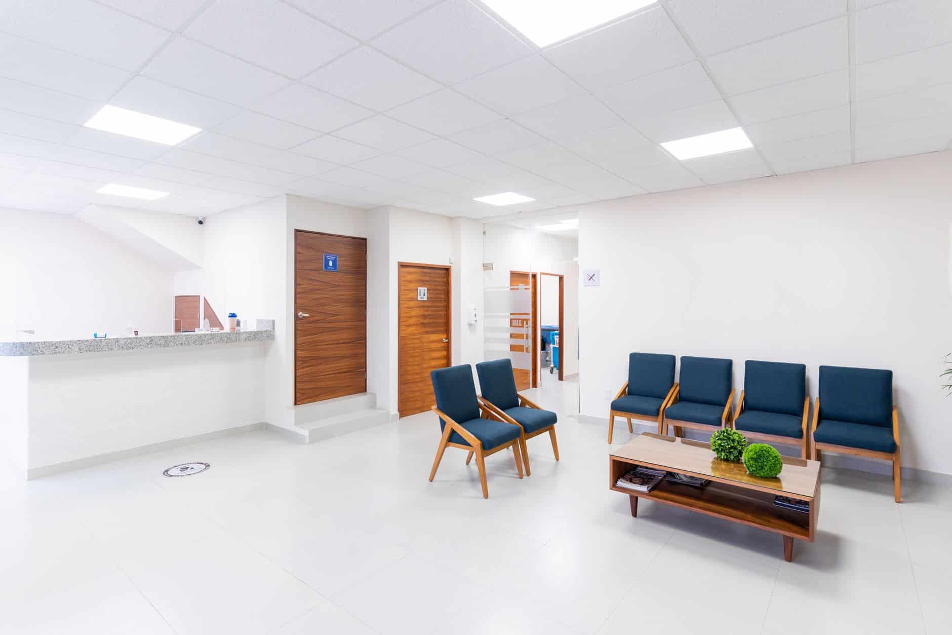 Una sala d'attesa luminosa di un ambulatorio, con il bancone dell'accoglienza, sedie blu e un tavolino con riviste. Spray psichedelico