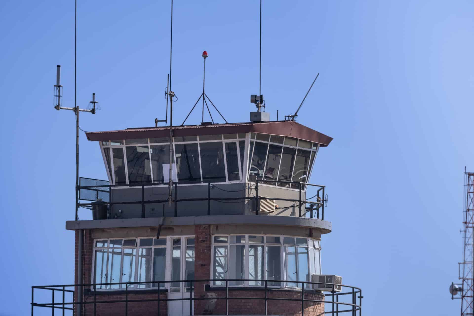 Torre di controllo di un aeroporto con vetrate, antenne e un faro rosso sul tetto, contro un cielo azzurro. Contrails